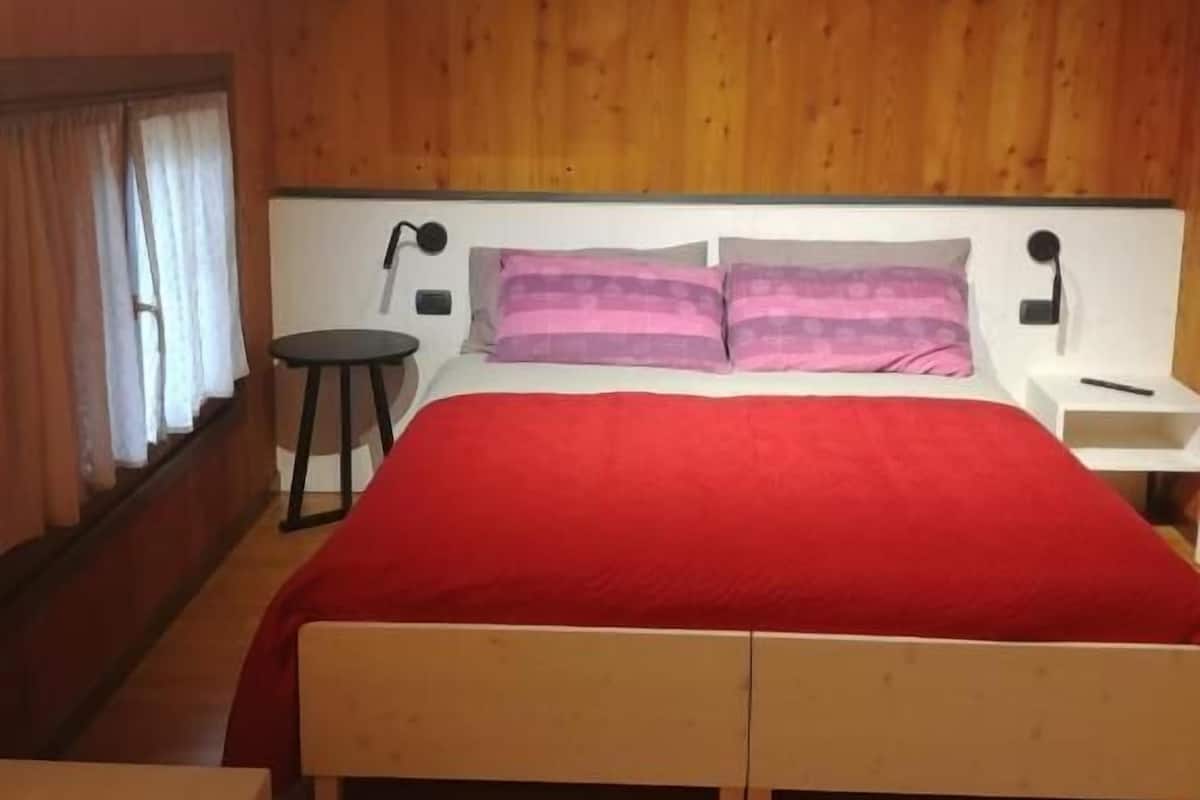 Ein Zimmer mit Holzwandverkleidung, einem Bett, einem Nachttisch, einem kleinen Tisch und einem Fenster mit Vorhängen.