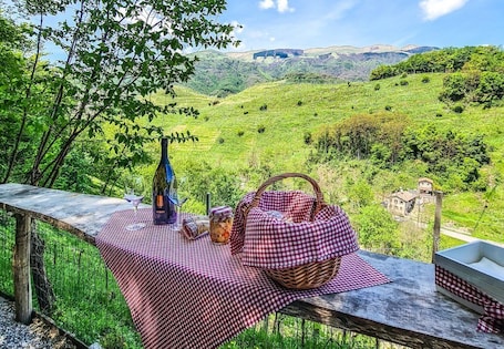 A picnic setup with a checkered tablecloth, a bottle, a basket, and a jar of food on a wooden table overlooking a lush valley.