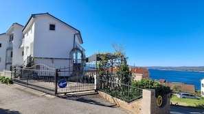 Ein weißes Gebäude mit einem von einem Tor gesäumten Eingang, einem blauem Pfeilschild und Blick auf das Meer.