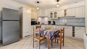 Una cocina con armarios blancos, un refrigerador de acero inoxidable, una mesa de comedor con sillas e iluminación colgante.