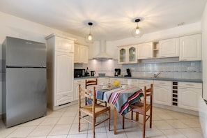 A kitchen with white cabinets, a stainless steel refrigerator, a dining table with chairs, and pendant lighting.