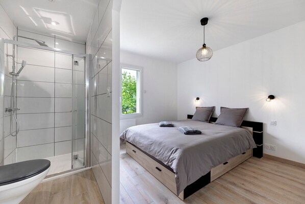 A modern bedroom with a glass shower enclosure, a wooden bed frame, and a large window providing natural light.
