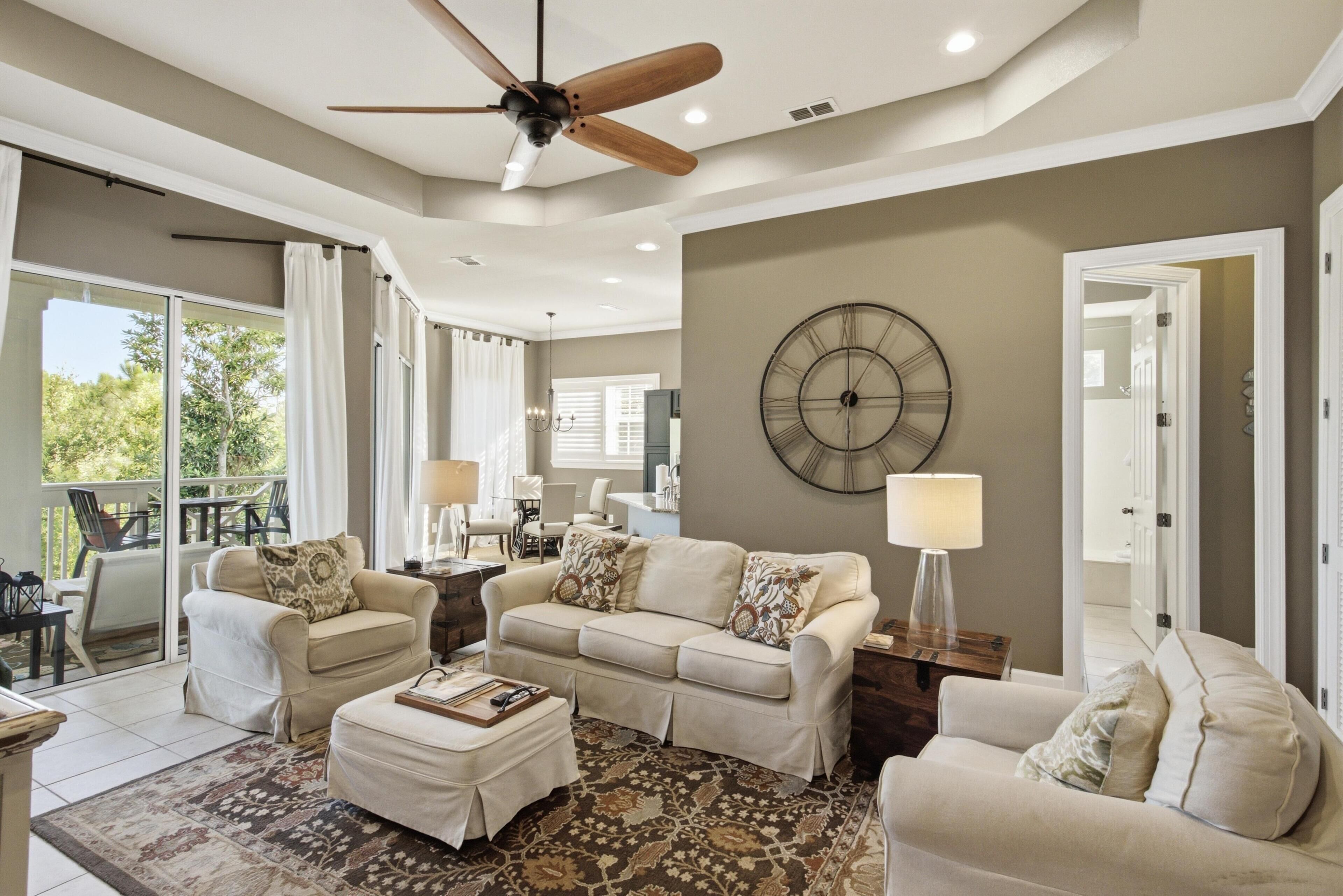 A living room with a ceiling fan, a large wall clock, and a view of the outdoors.
