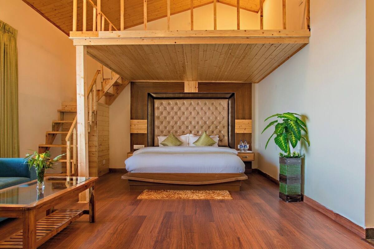 Ein Schlafzimmer mit Holzbalkendecke, einem gepolsterten Kopfteil, einem Bett mit Kissen, einem Nachttisch, einem Glastisch, einem Sofa, einer Treppe und einer Zimmerpflanze.