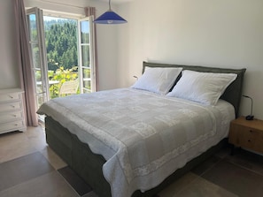 Un dormitorio con cama, mesita de noche, cómoda y vistas al verdor por una ventana.