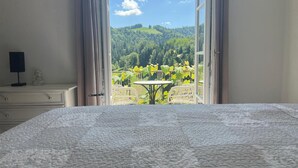 Quarto com uma cama grande, mesa de cabeceira com luminária e vista para uma varanda com mesa e cadeiras.