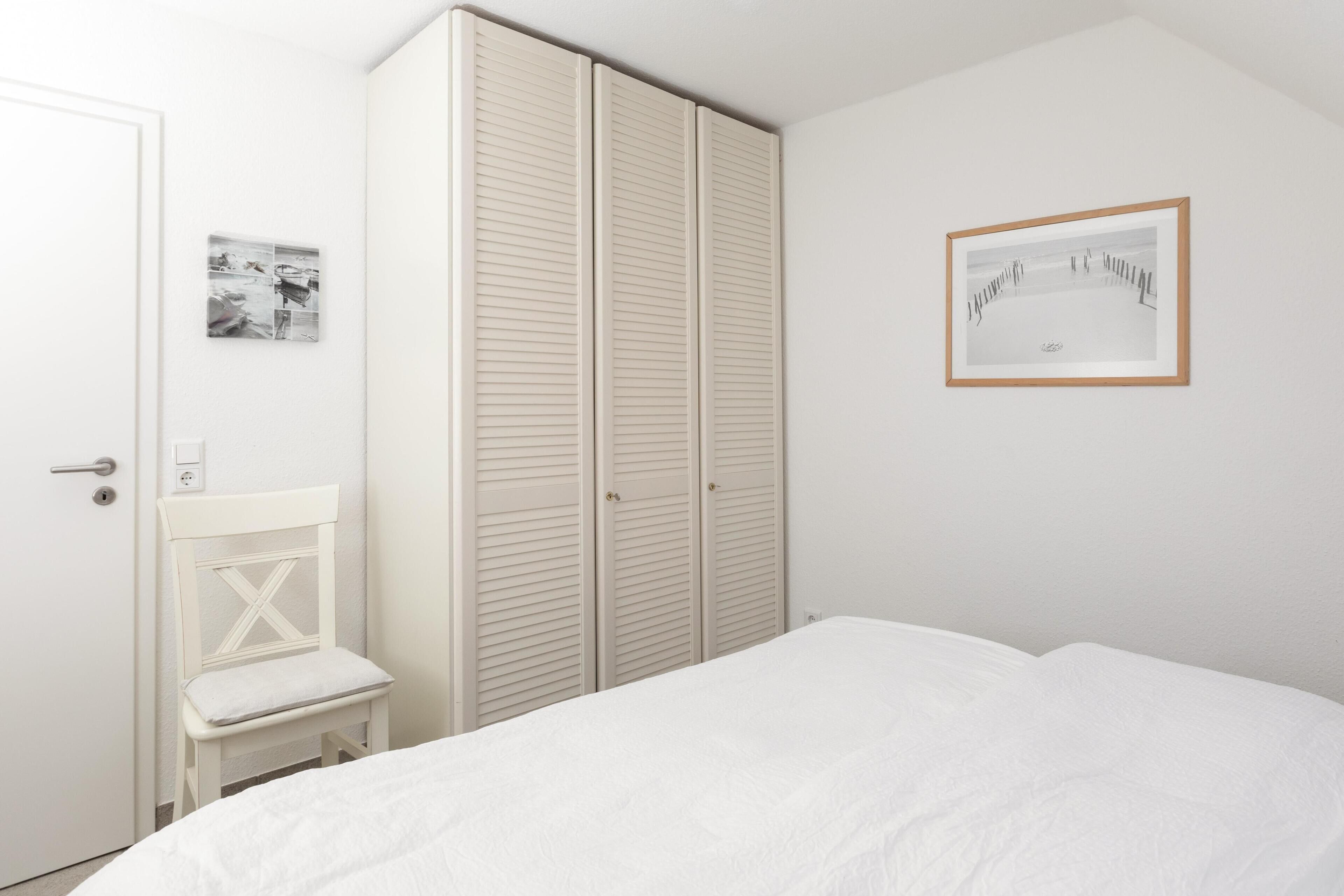 Une chambre à coucher avec un lit, une chaise blanche, une armoire et un tableau encadré au mur.