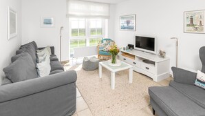 Un salon moderne avec un canapé gris, une table basse blanche ornée d’un vase, un téléviseur à écran plat et une grande fenêtre donnant sur la campagne.