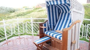A wicker lounge chair with a striped cushion, a small wooden table, and a view of a grassy landscape with a white fence. - "Dünenblick" at Haus Sanderling - your vacation paradise with a view (Juist)