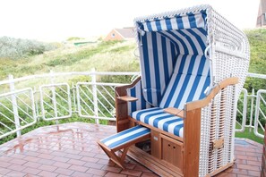 A wicker lounge chair with a striped cushion, a small wooden table, and a view of a grassy landscape with a white fence. - "Dünenblick" at Haus Sanderling - your vacation paradise with a view (Juist)