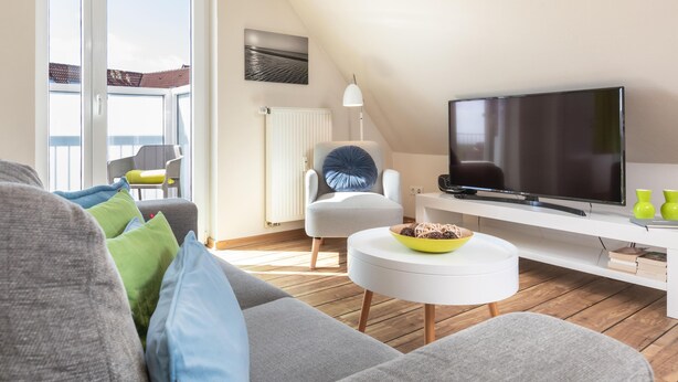 Un salon moderne avec un canapé, un téléviseur, une petite table d’appoint blanche et une vue sur l’extérieur.