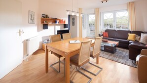 Een moderne woonkamer met een eettafel, stoelen, een bank en een salontafel. Er is een raam met gordijnen, een radiator en een plank met boeken en decoratieve objecten.
