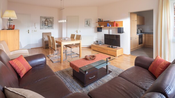 Een woonkamer met een bruine leren bank, een glazen salontafel, een televisie en een eethoek met een houten tafel en stoelen.