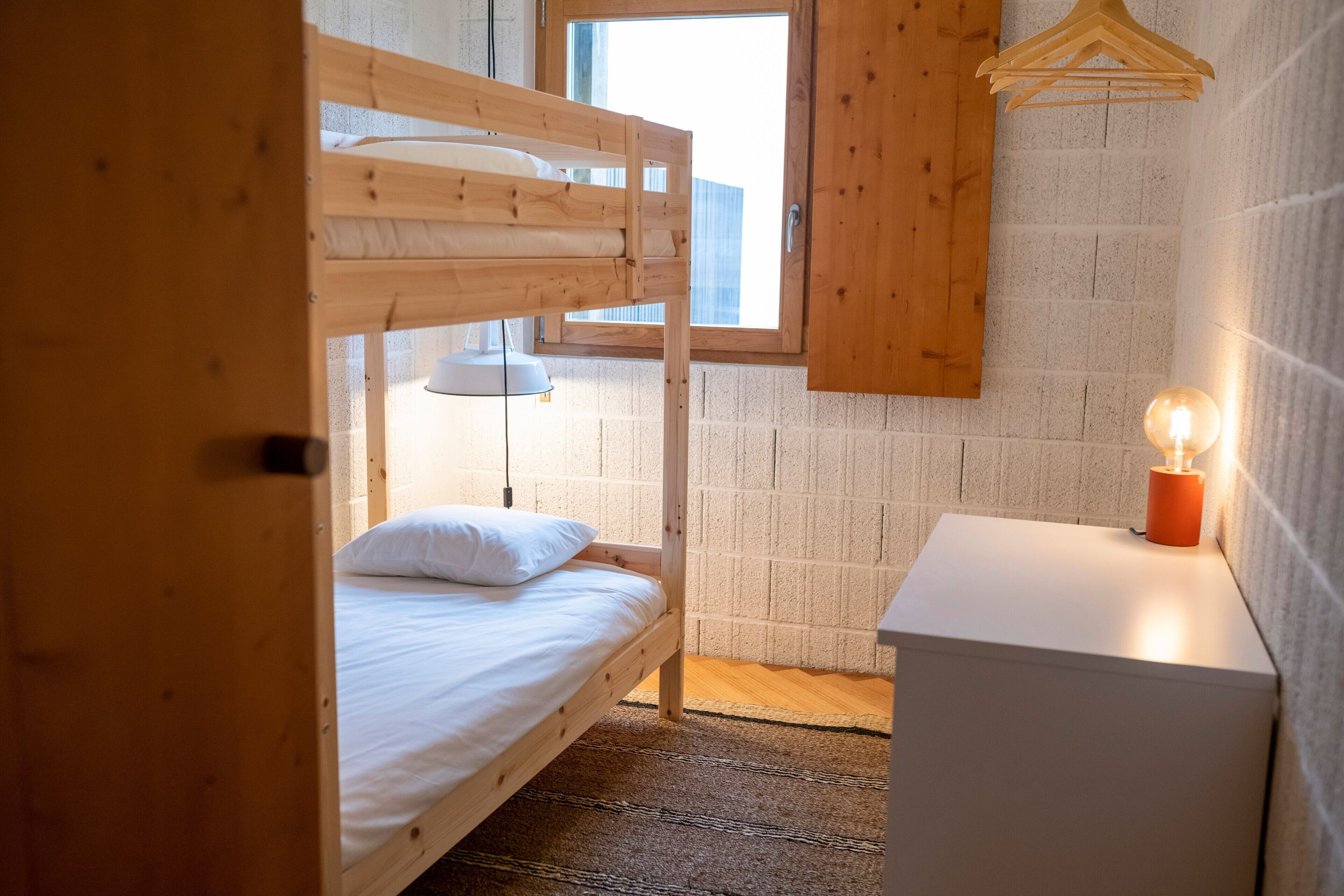 Una cama litera con un perchero de madera colgante, una mesita de noche con una vela encendida y una ventana con vistas al exterior.