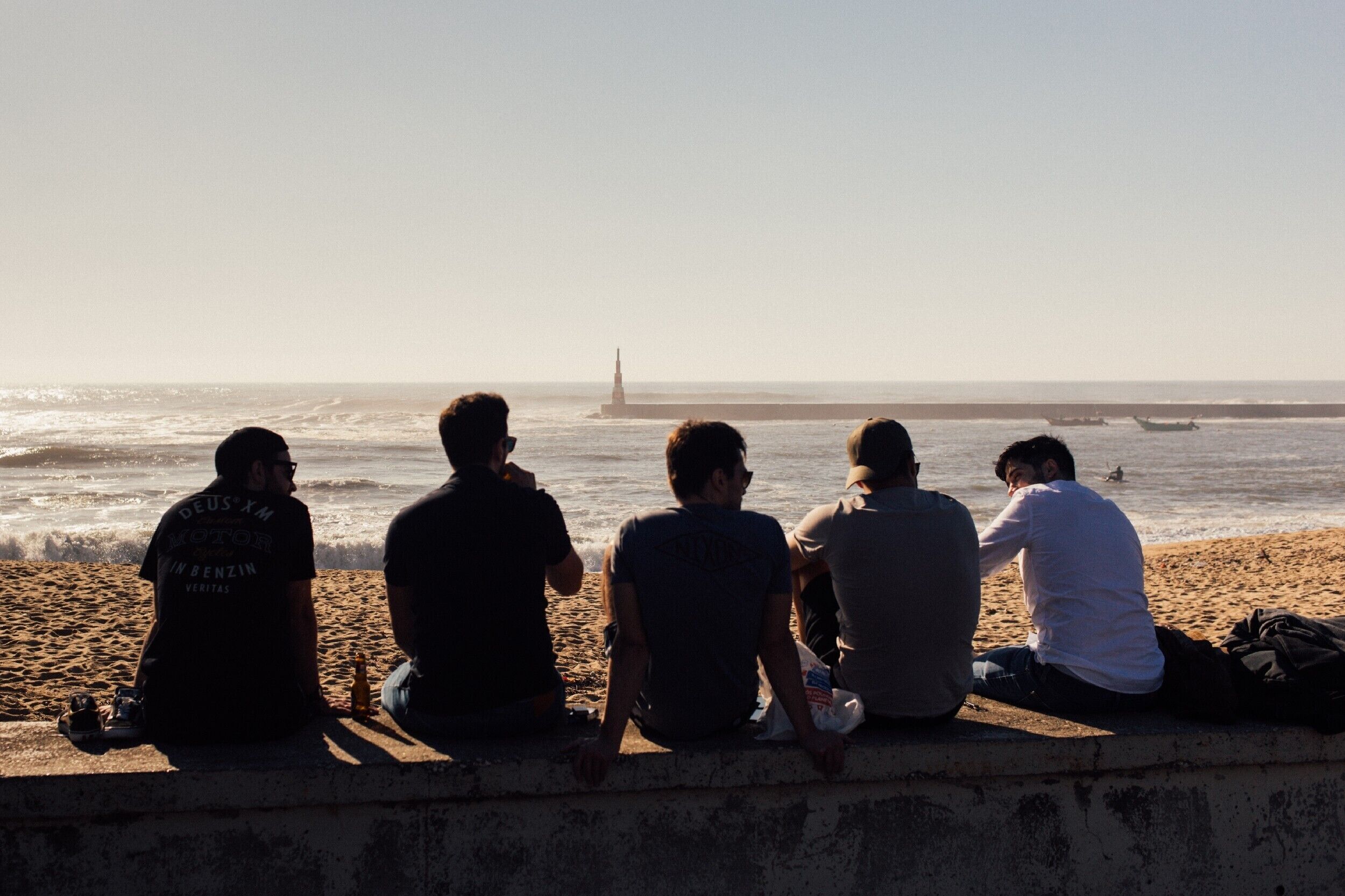 Eine Gruppe von Menschen sitzt auf einer Betonkante am Meer und blickt auf den Ozean sowie einen fernen Leuchtturm.