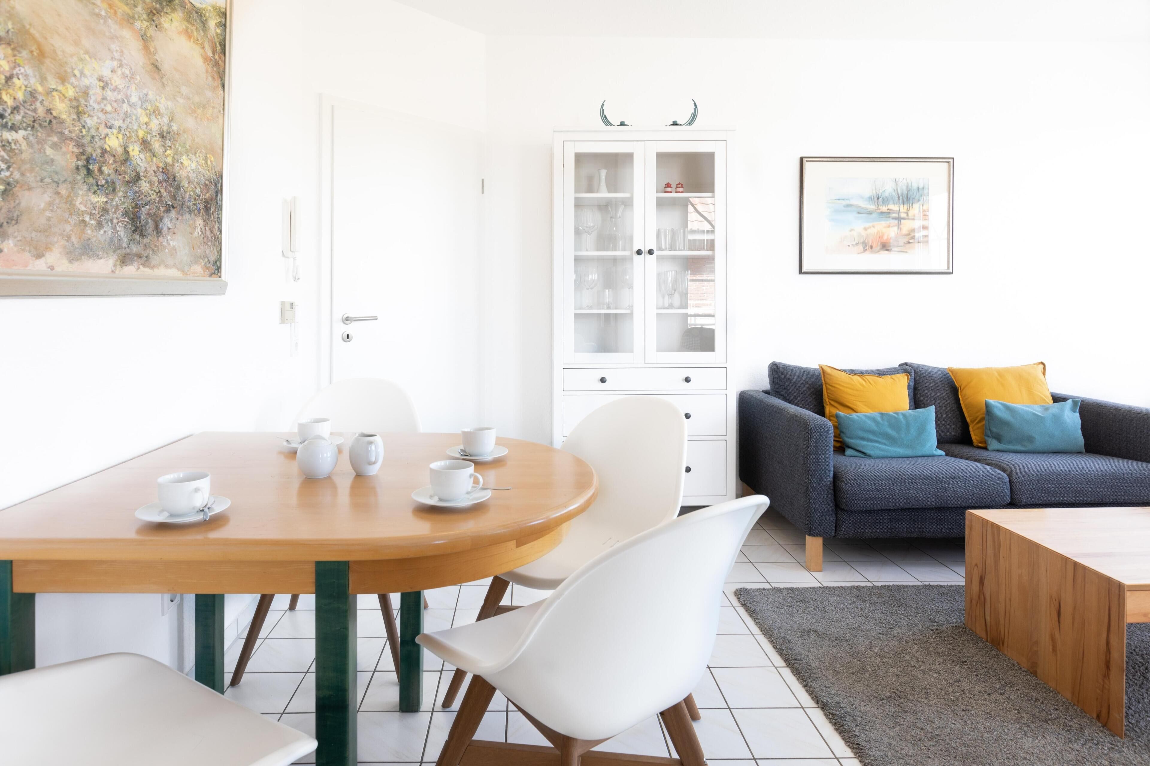 A modern living room with a dining table, white chairs, a gray sofa with yellow and blue cushions, a cabinet with glass doors, and a painting on the wall.