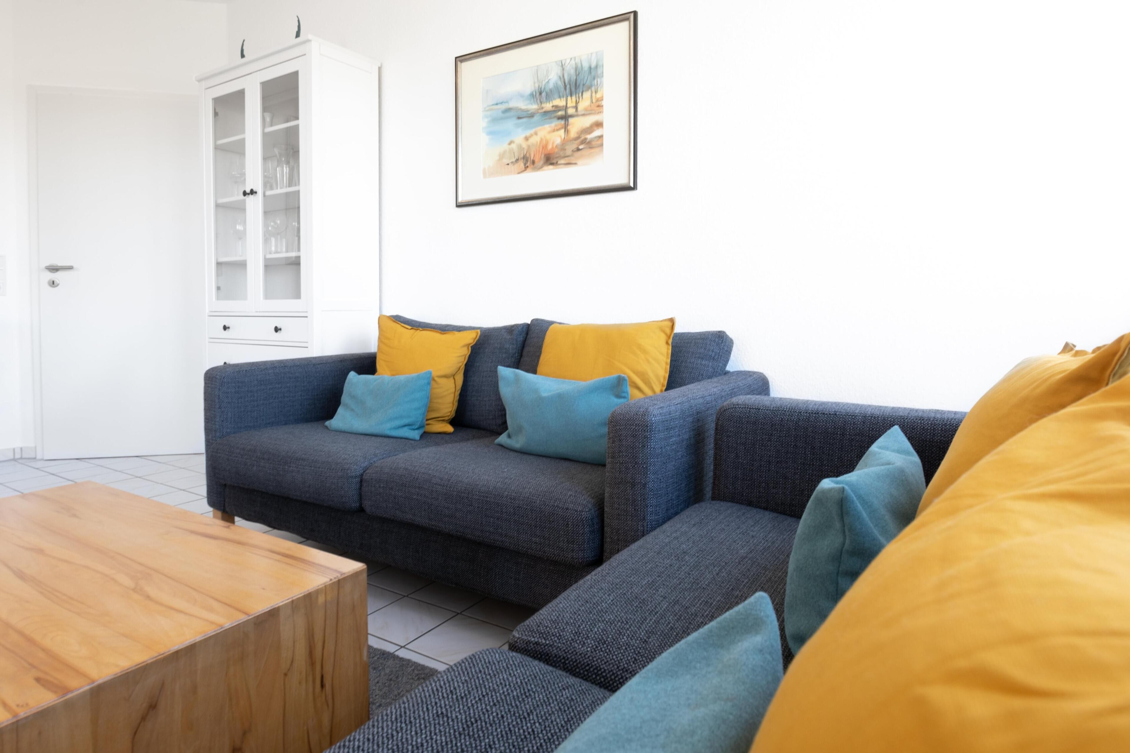 Un salón con un sofá gris, cojines amarillos y azules, una mesa de centro de madera y un aparador blanco con puertas de cristal.