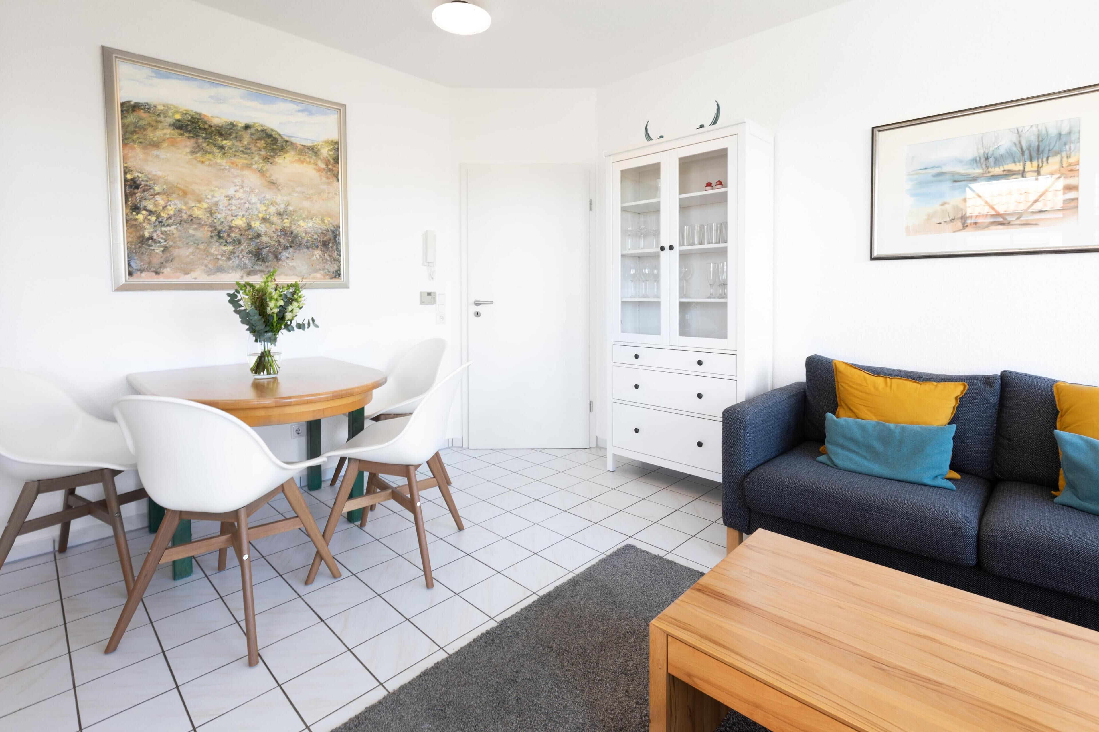 Een moderne woonkamer met een eettafel, witte stoelen, een bank met kussens en een kast met glazen.