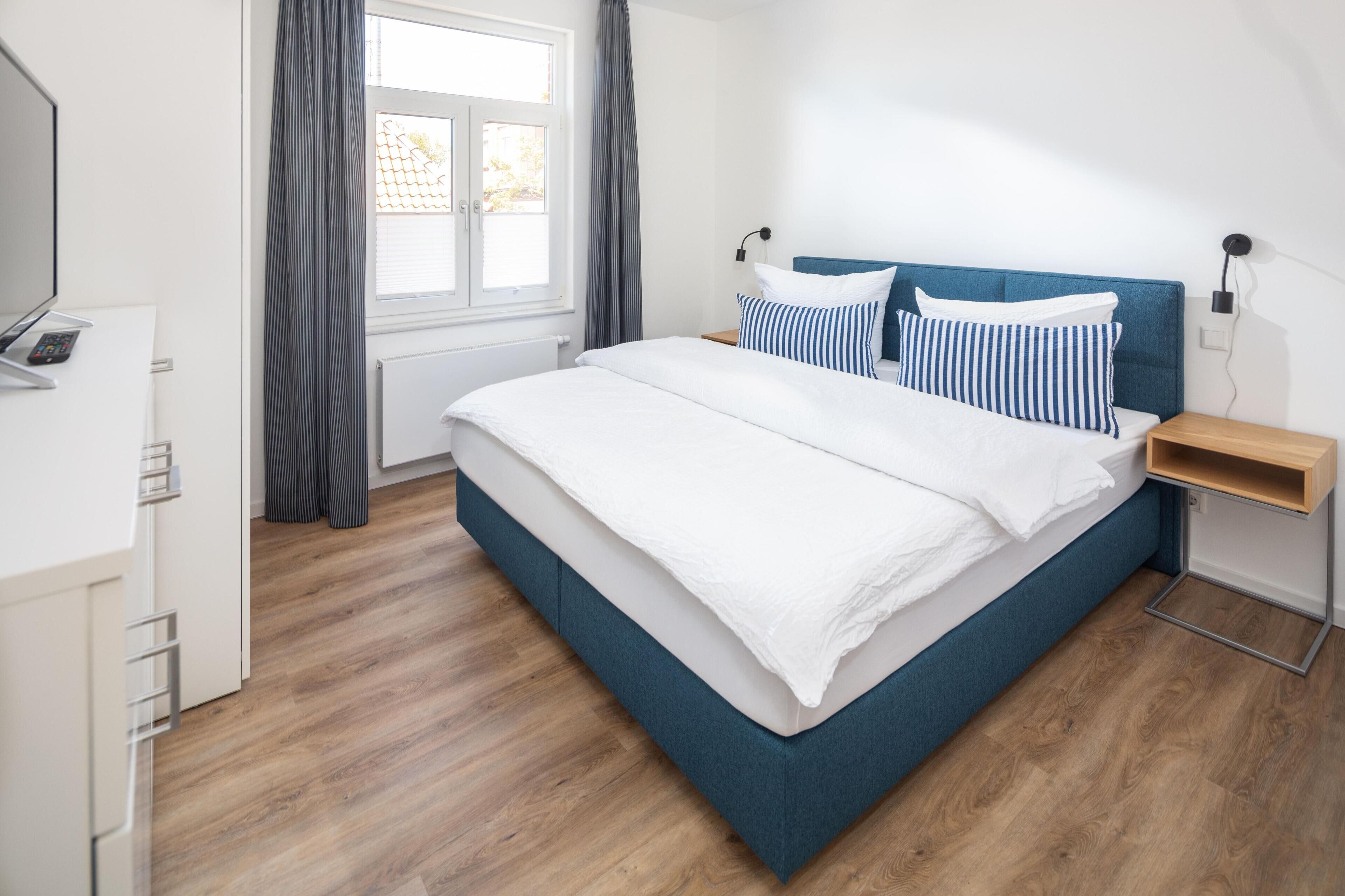 Un dormitorio con cama, televisor, ventana con cortinas y piso de madera.