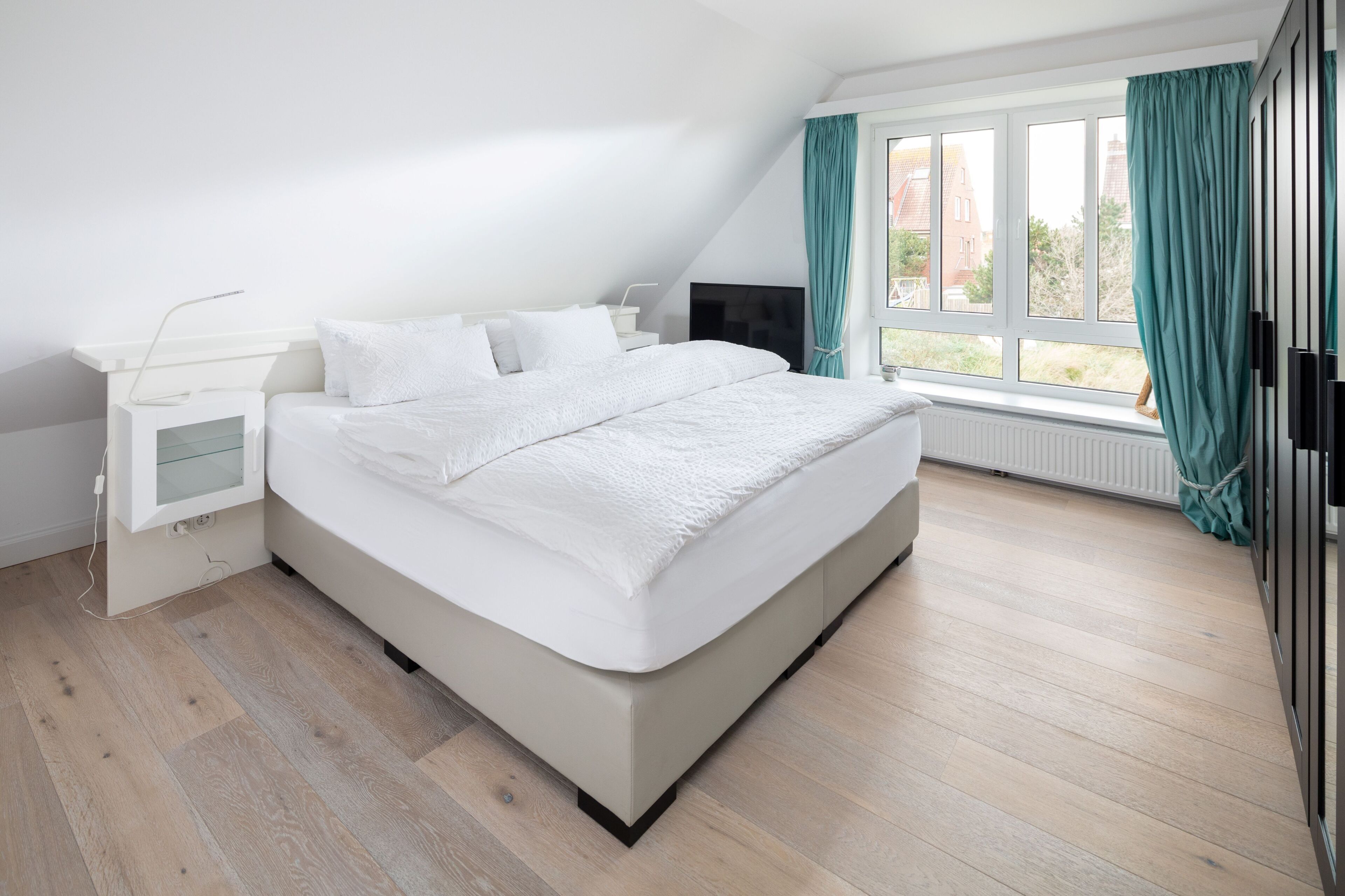 A bedroom with a bed, a bedside table, a TV, and a window with curtains.