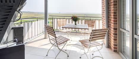 A balcony with a grill, two wooden chairs, a table with a vase, and a view of the landscape.