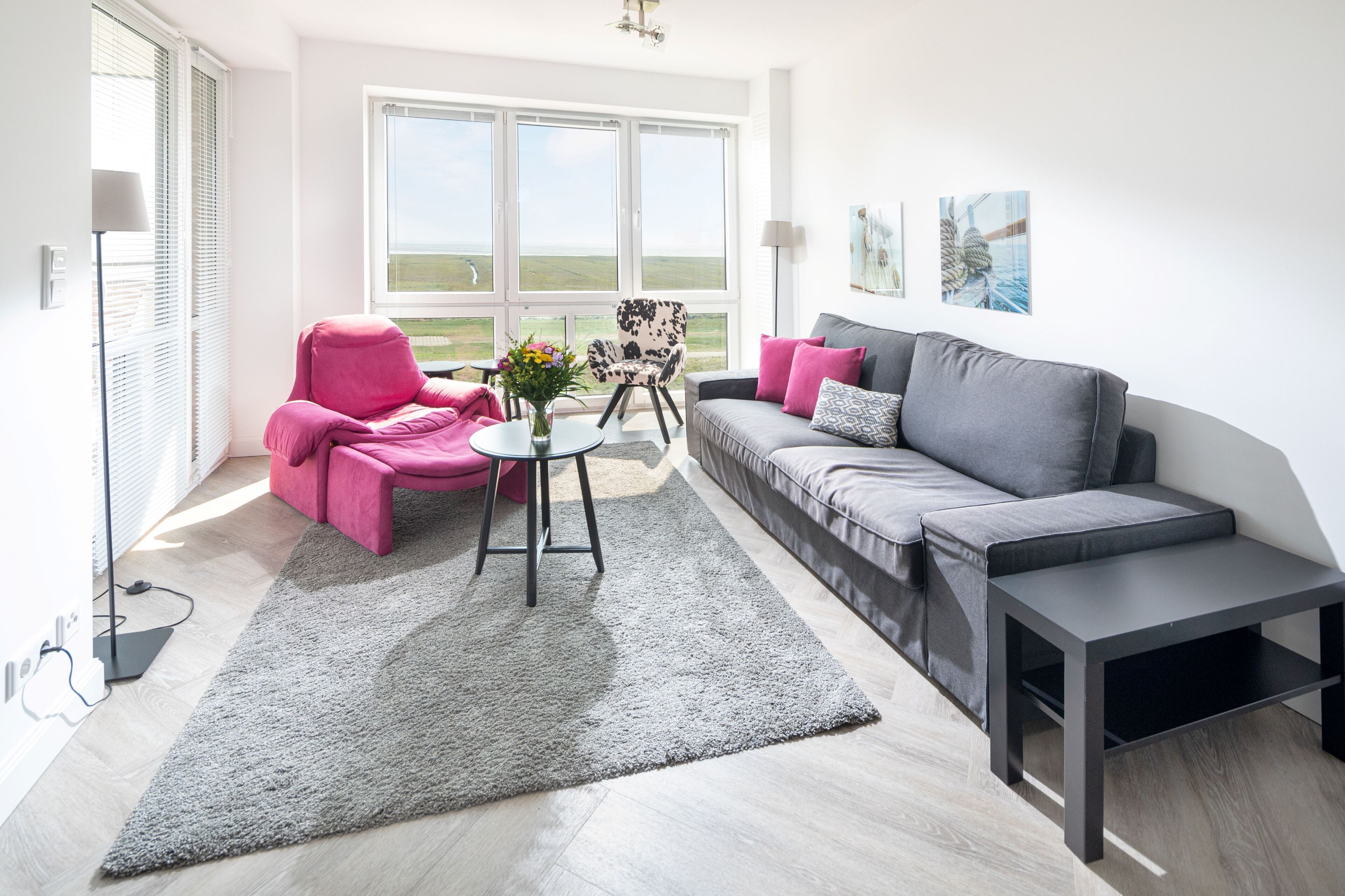 Un salon avec un canapé gris, un fauteuil rose, une table basse et une vue sur l’extérieur.