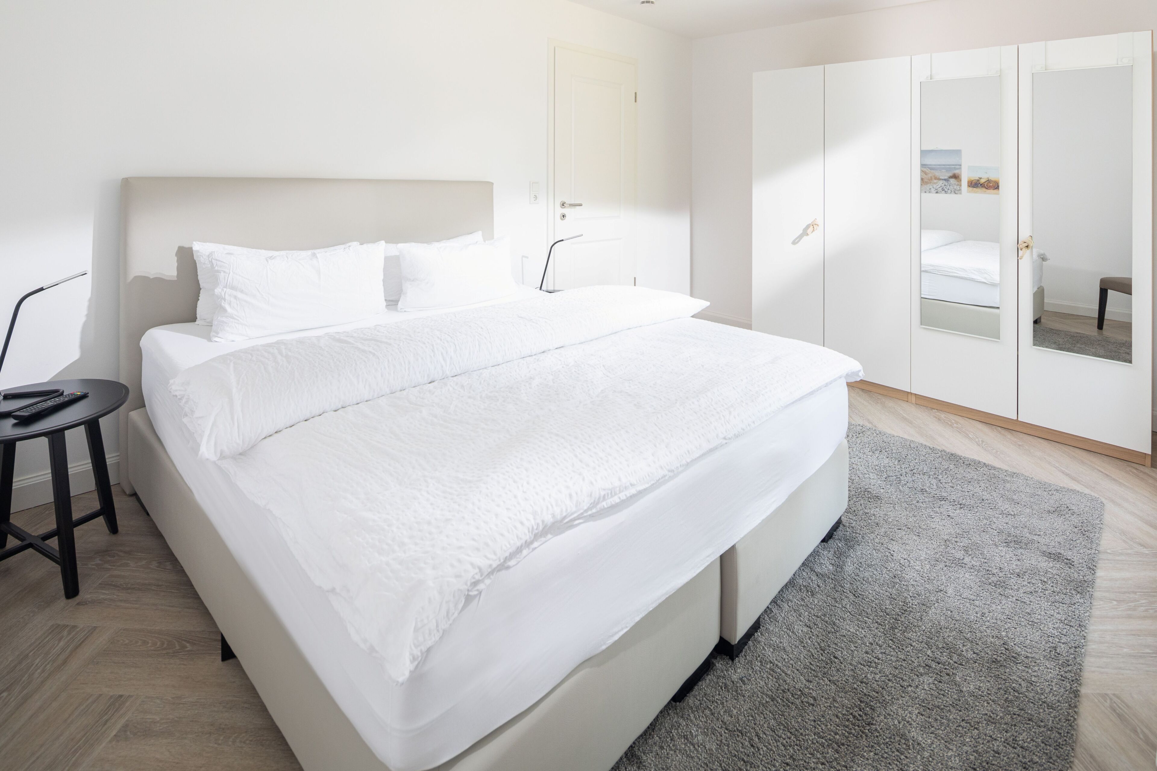 A modern bedroom with a large bed, a bedside table, a wardrobe, and a mirror.
