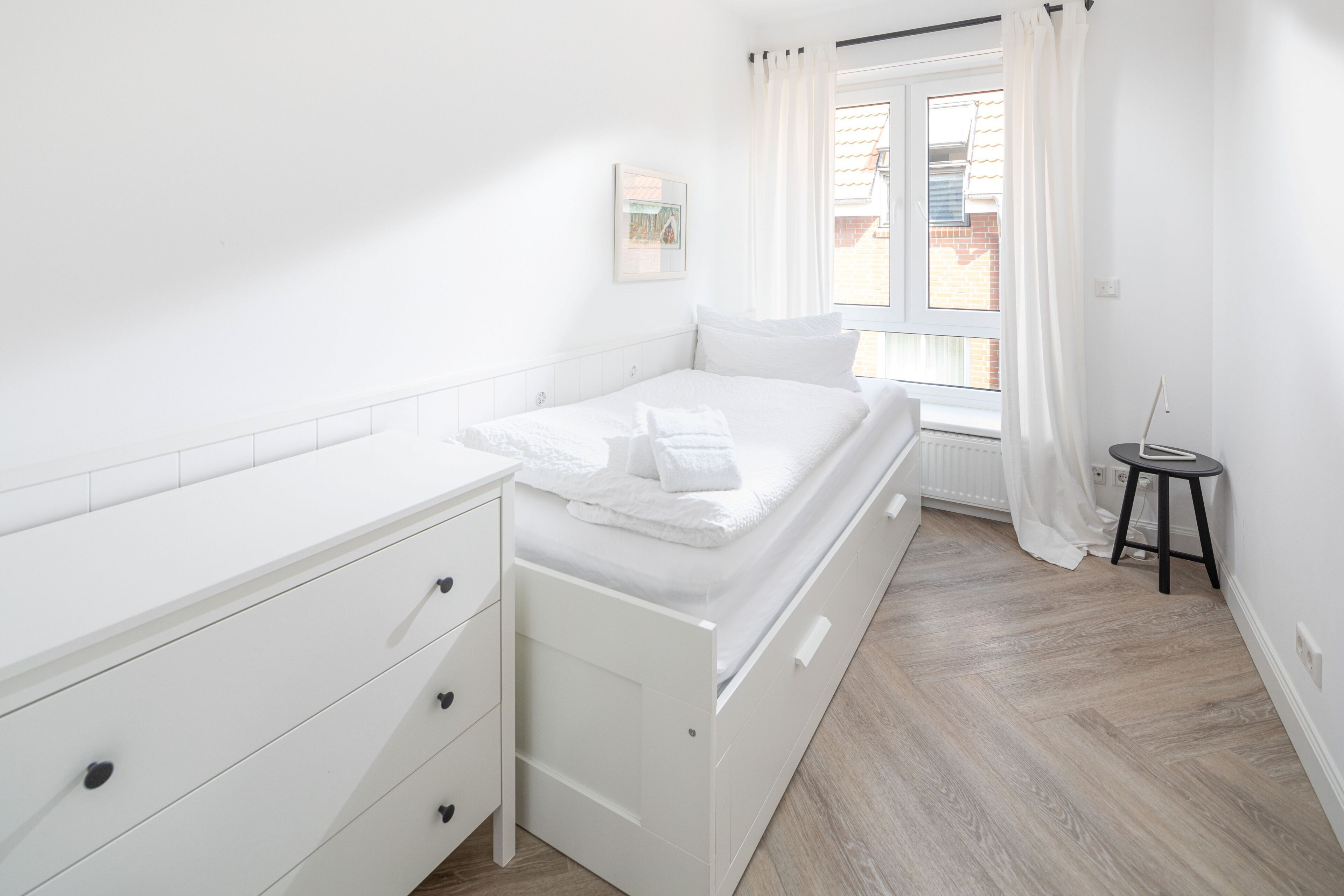 A bedroom with a bed, a white dresser, a small table, and a window with curtains.