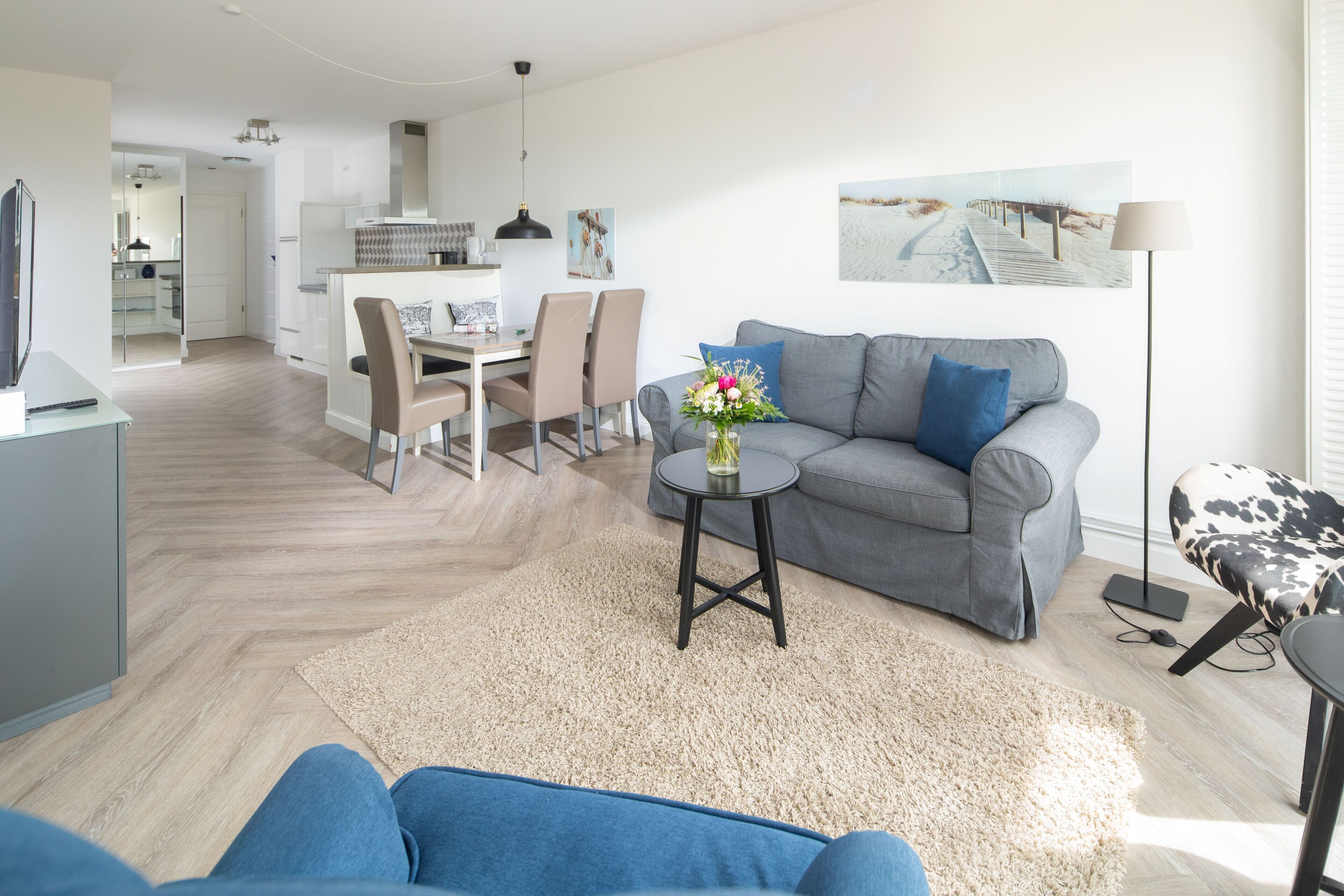 A modern living room with a grey sofa, a small round table with a vase, and a dining area in the background.