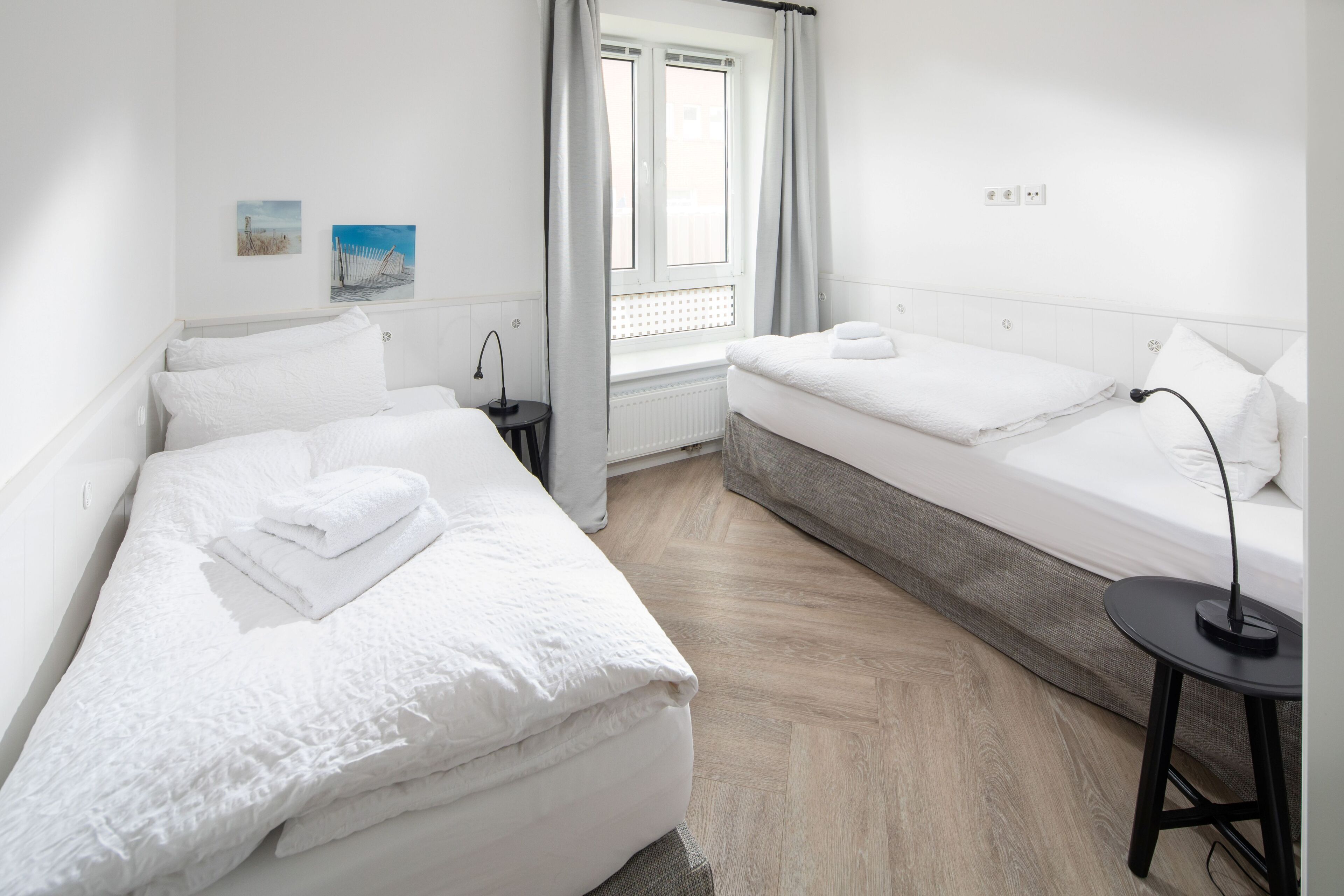 Ein Hotelzimmer mit zwei Betten, einem Fenster mit Vorhängen, einem kleinen Tisch und einer wandmontierten Steckdose.