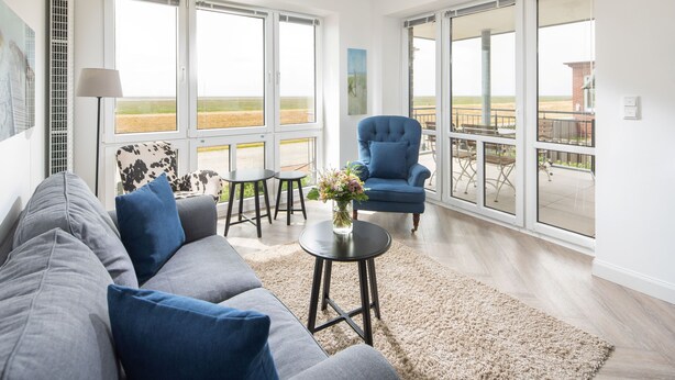Una sala de estar moderna con un sofá, un sillón azul, una mesita con un florero y amplias ventanas que ofrecen vistas al exterior.