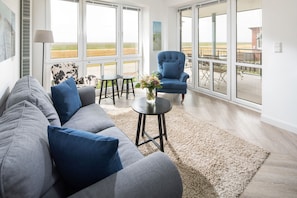 A modern living room with a sofa, a blue armchair, a small table with a vase, and large windows offering a view of the outdoors. - Relaxation and Adventure: Your Dream Vacation at the Sonnenrose Vacation Rental! (Juist)