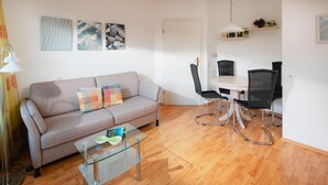 A modern living room with a sofa, glass coffee table, dining table with chairs, and artwork on the wall. - "Experience an unforgettable vacation at Haus Sanderling Kleeblatt!" (Juist)
