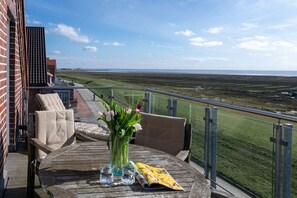 A balcony with a view of a flat landscape, a table with a vase of flowers, and two chairs. - Cozy vacation apartment with a spectacular ocean view (Juist)
