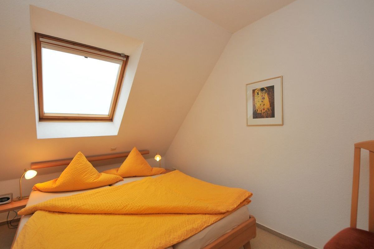 Une chambre avec une lucarne, un lit avec une literie jaune, une lampe de chevet et un tableau encadré au mur.