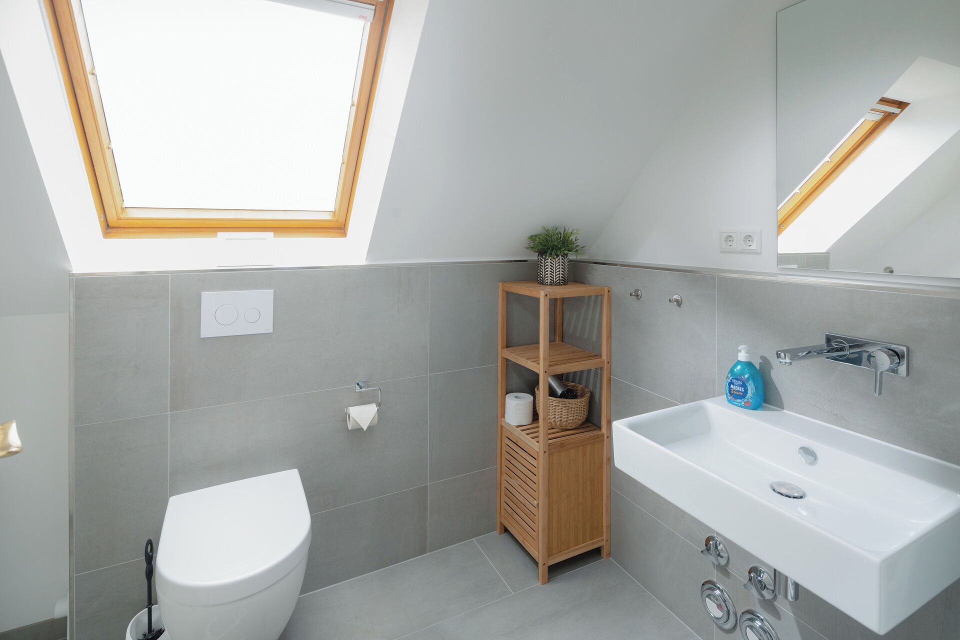 A modern bathroom with a skylight, a white toilet, a wooden shelf with toiletries, and a white sink with a mirror.