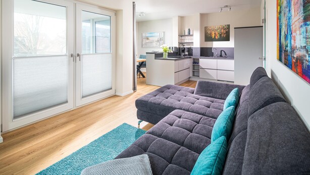 Een moderne woonkamer met een grijze bank, een keuken op de achtergrond en een groot raam met zonwering.