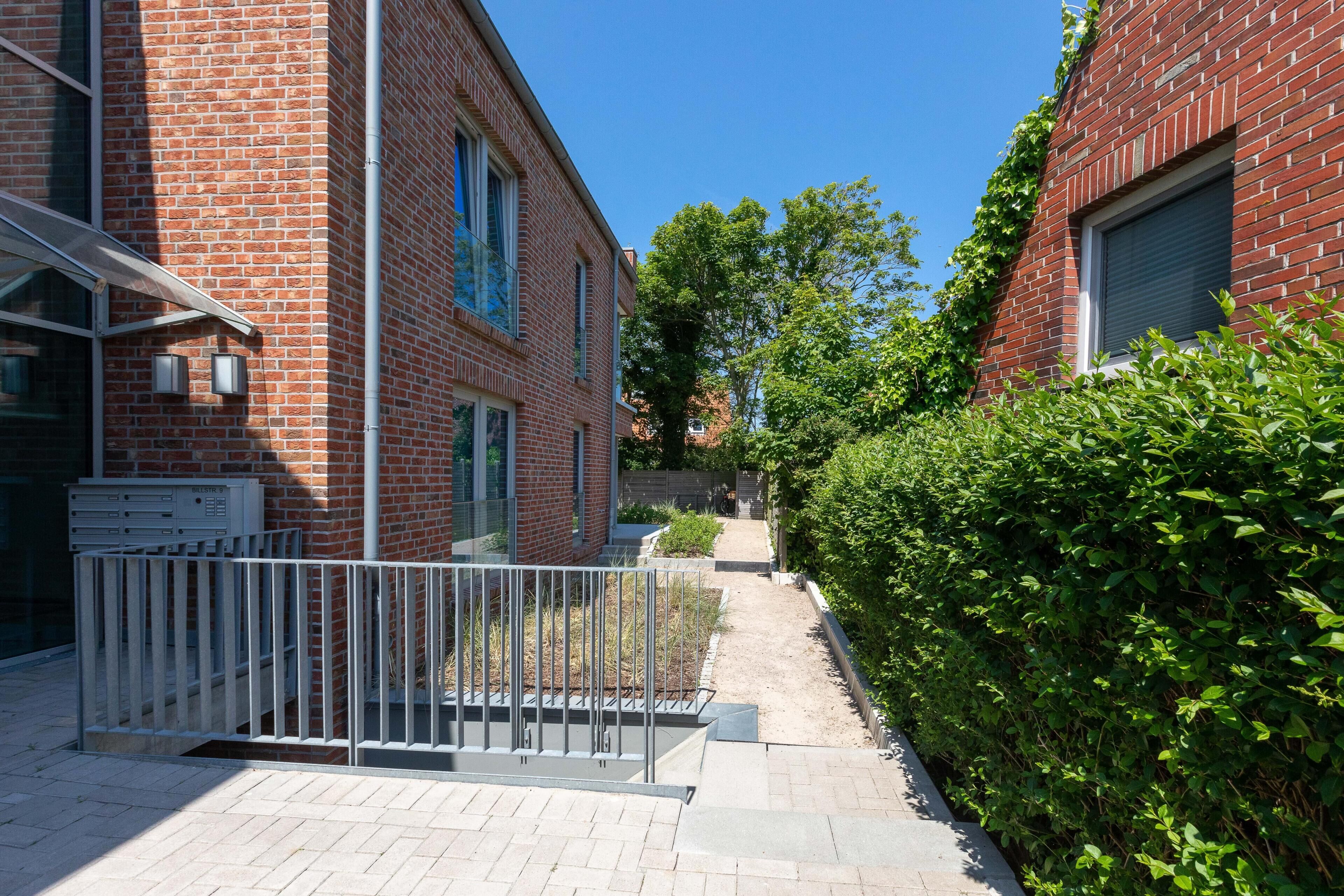 A brick building with a metal fence, a paved walkway, and green foliage.