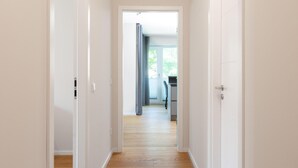 Een gang met houten vloer, witte muren en deuren die leiden naar een kamer met een raam en bureau.