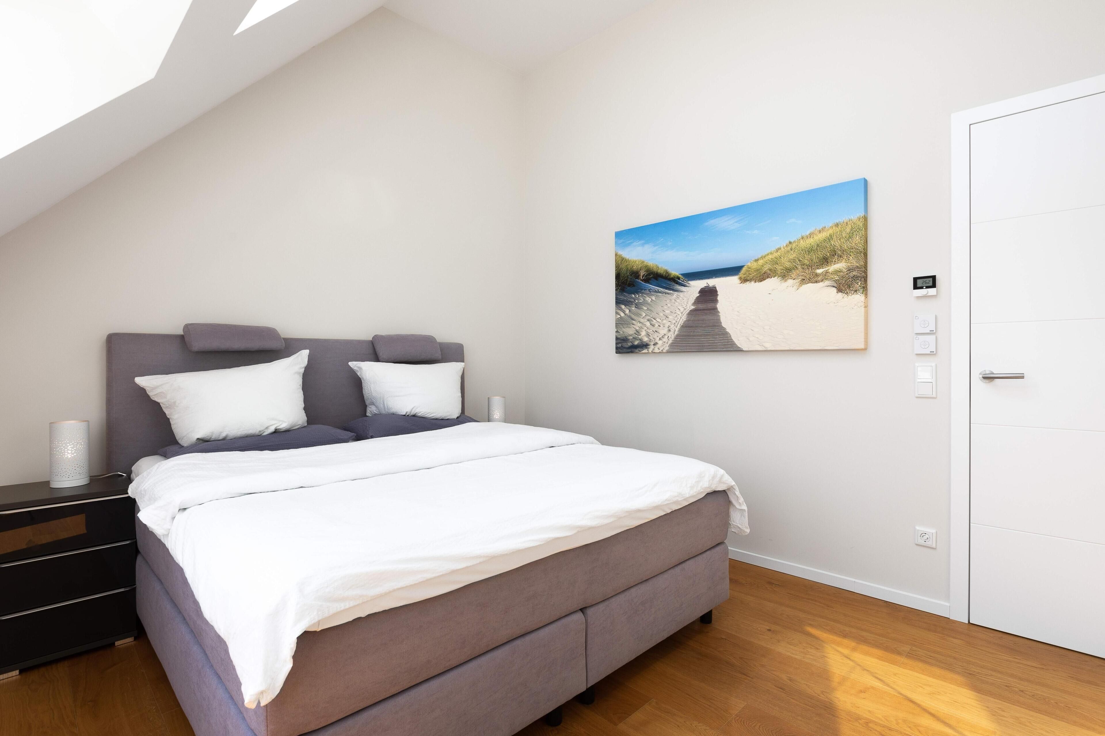 Une chambre à coucher comprenant un lit, une table de chevet, un thermostat fixé au mur et un tableau sur le thème de la plage.