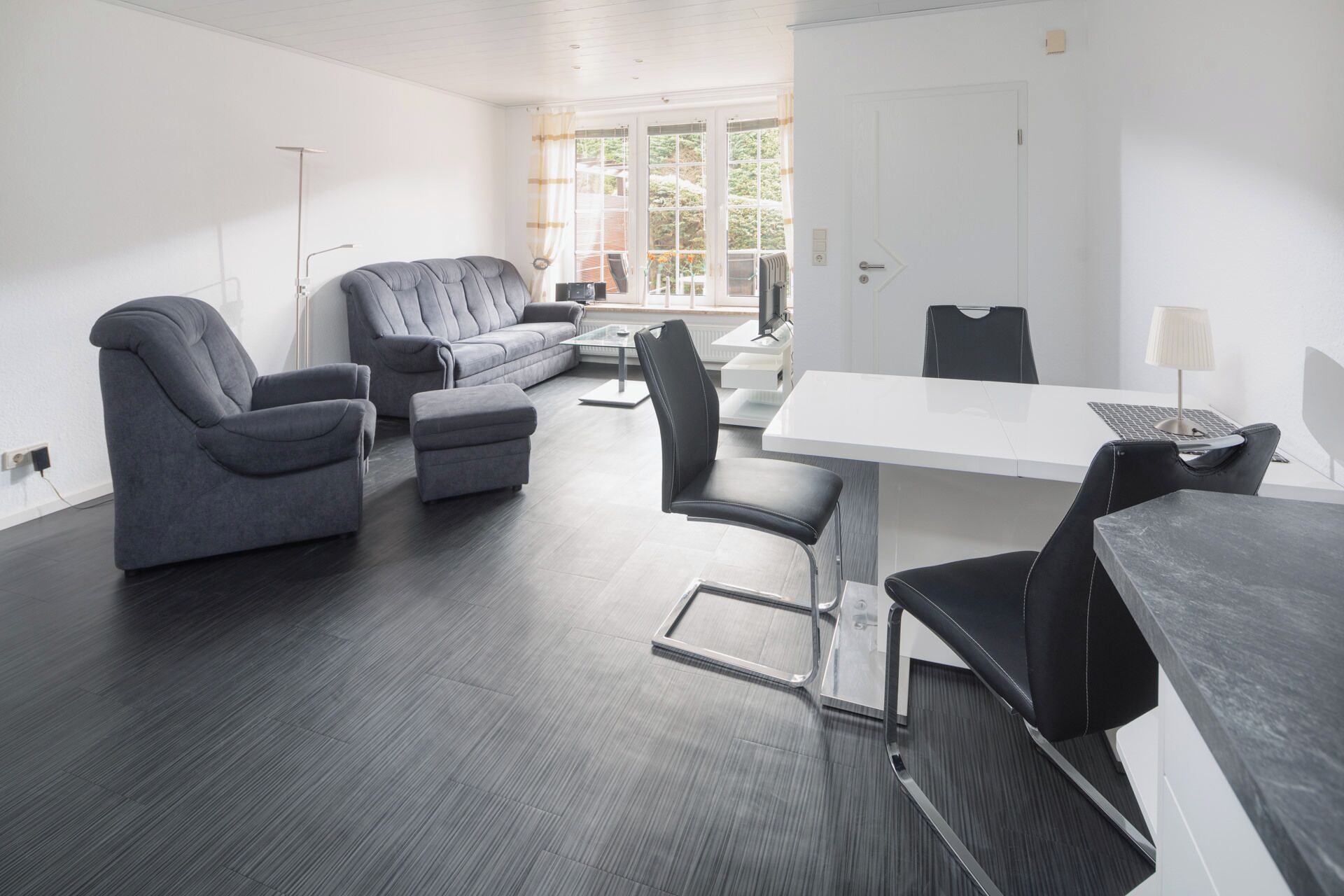 Ein modernes Wohnzimmer mit einem grauen Sofa, passenden Sesseln, einem Esstisch und Blick durch große Fenster.