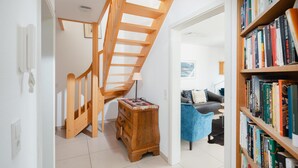 Eine Treppe mit Holzvertäfelung, eine kleine Holzkommode, eine Lampe, eine Bücherwand voller Bücher und ein blauer Stuhl.