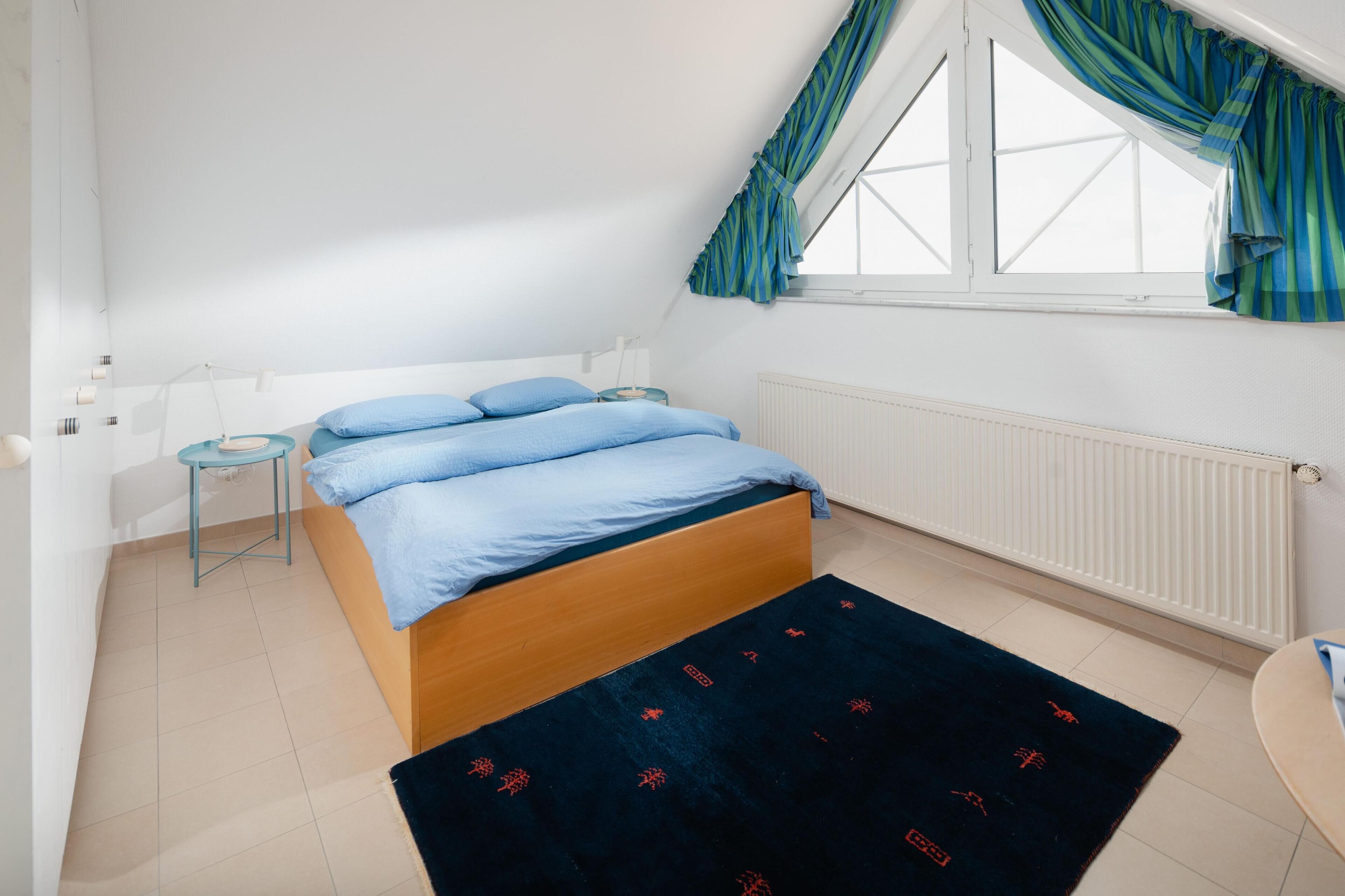 Ein Schlafzimmer mit einem Bett, einem kleinen Nachttisch, einem Heizkörper und einem Fenster mit Vorhängen.