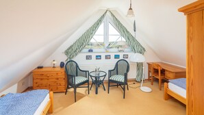 Een knus zolderkamer met schuine wanden, een houten dressoir, twee stoelen, een klein rond tafeltje, een lamp en een bed.