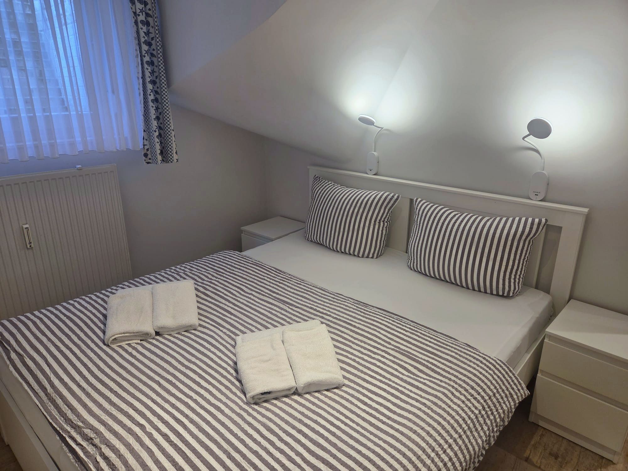 Cama bem arrumada com roupa de cama listrada e duas almofadas, mesa de cabeceira com abajur e janela com cortinas.