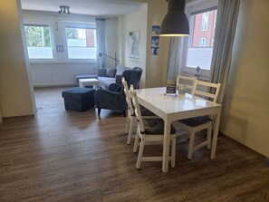 A dining area with a white table and chairs, a small white side table, and a window with curtains. - "Idyllic vacation apartment Seute Deern Kajüte - your perfect retreat on the (Juist)