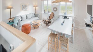 Ein modernes Wohnzimmer mit einer Couch, einem Couchtisch, Korbstühlen, einem Esstisch mit Barhocker, einem Fernseher und einem großen Fenster mit Blick.