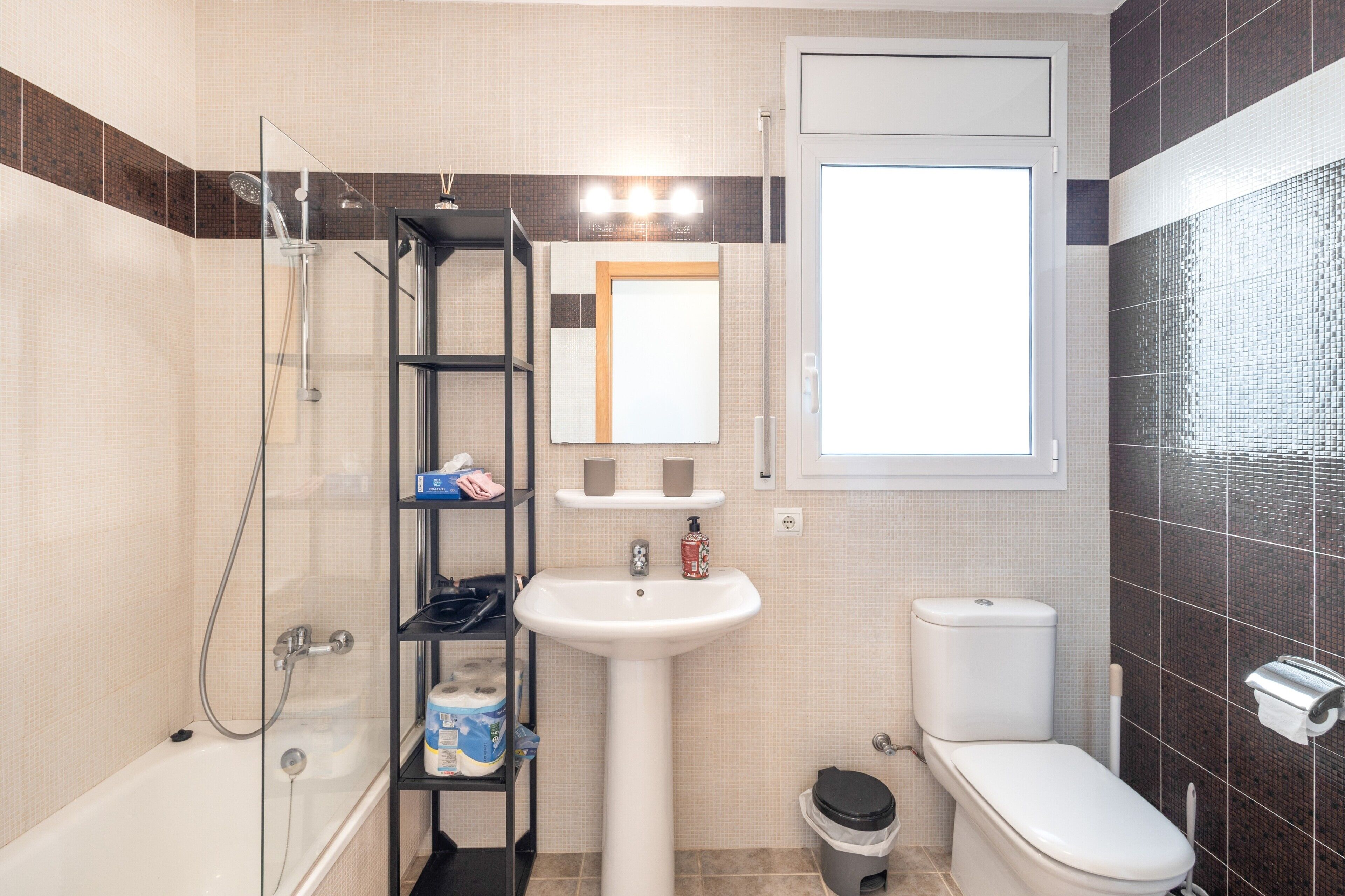 Un bany amb plat de dutxa de vidre, un lavabo blanc, un vàter i una estant amb productes de tocador.