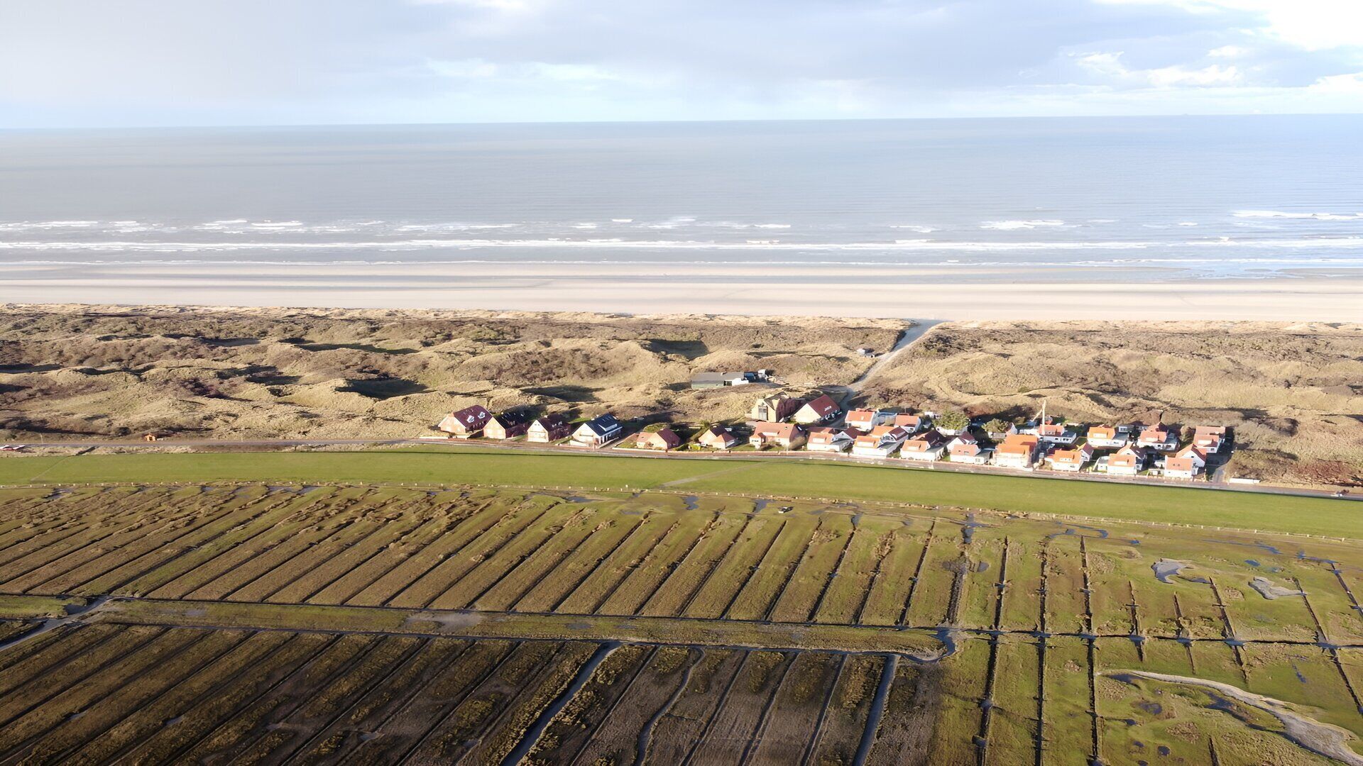 An aerial view of a coastal area with a beach, dunes, and a row of houses with a view of the sea.