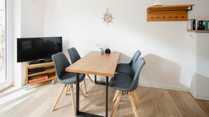 Ruokailutilassa on puinen pöytä, neljä harmaata tuolia, jalustalla oleva televisio ja kirjahylly, jossa on kirjoja.